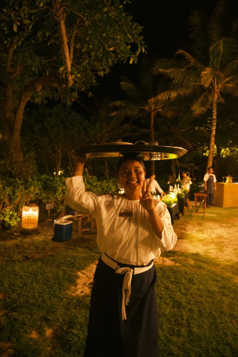 A smiling server carrying a tray overhead beneath palm trees at night